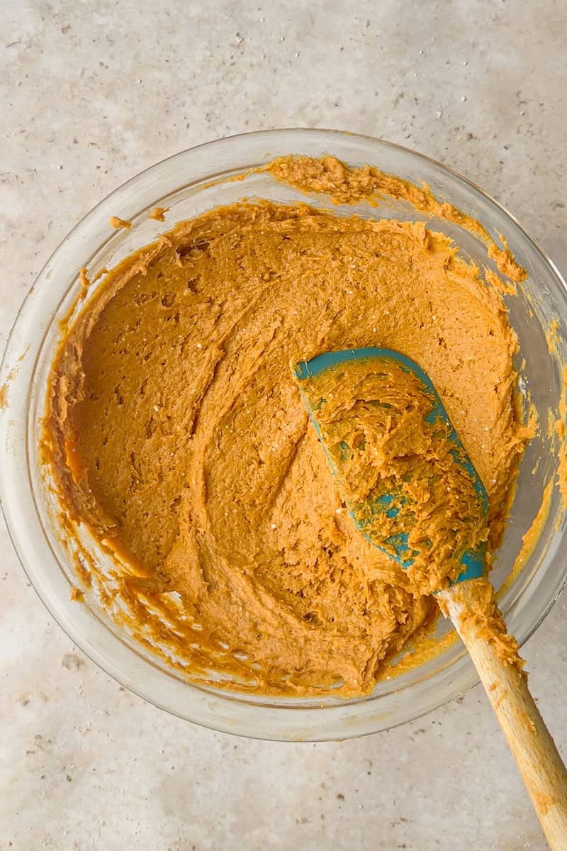 A glass bowl of thick pumpkin batter with a blue spatula resting in the mixture.