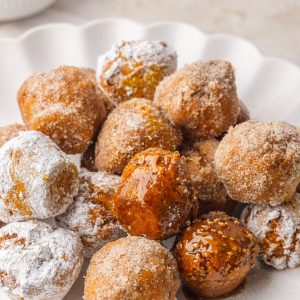 A pile of pumpkin donut holes on a white plate coated in cinnamon sugar, powdered sugar, and maple glaze.