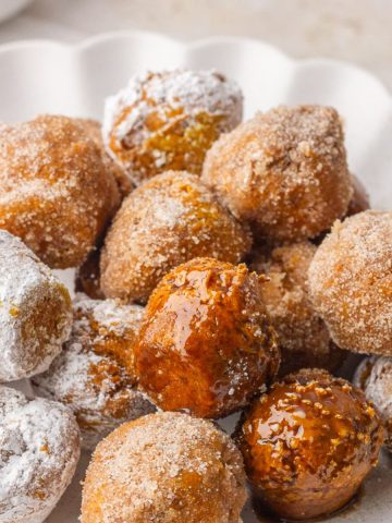 A pile of pumpkin donut holes on a white plate coated in cinnamon sugar, powdered sugar, and maple glaze.