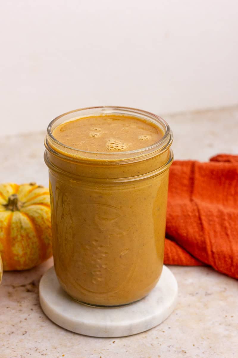A glass jar filled with homemade pumpkin spice sauce sitting on a marble coaster next to a small pumpkin and an orange towel.