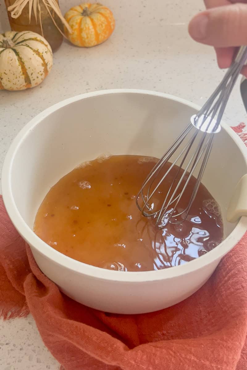 A hand whisking brown sugar and water together in a white saucepan to start the sauce.