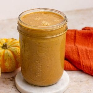 A close-up of a jar filled with creamy pumpkin spice sauce with a pumpkin and orange towel beside it.
