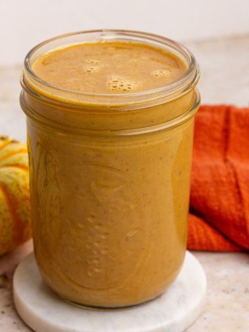 A close-up of a jar filled with creamy pumpkin spice sauce with a pumpkin and orange towel beside it.