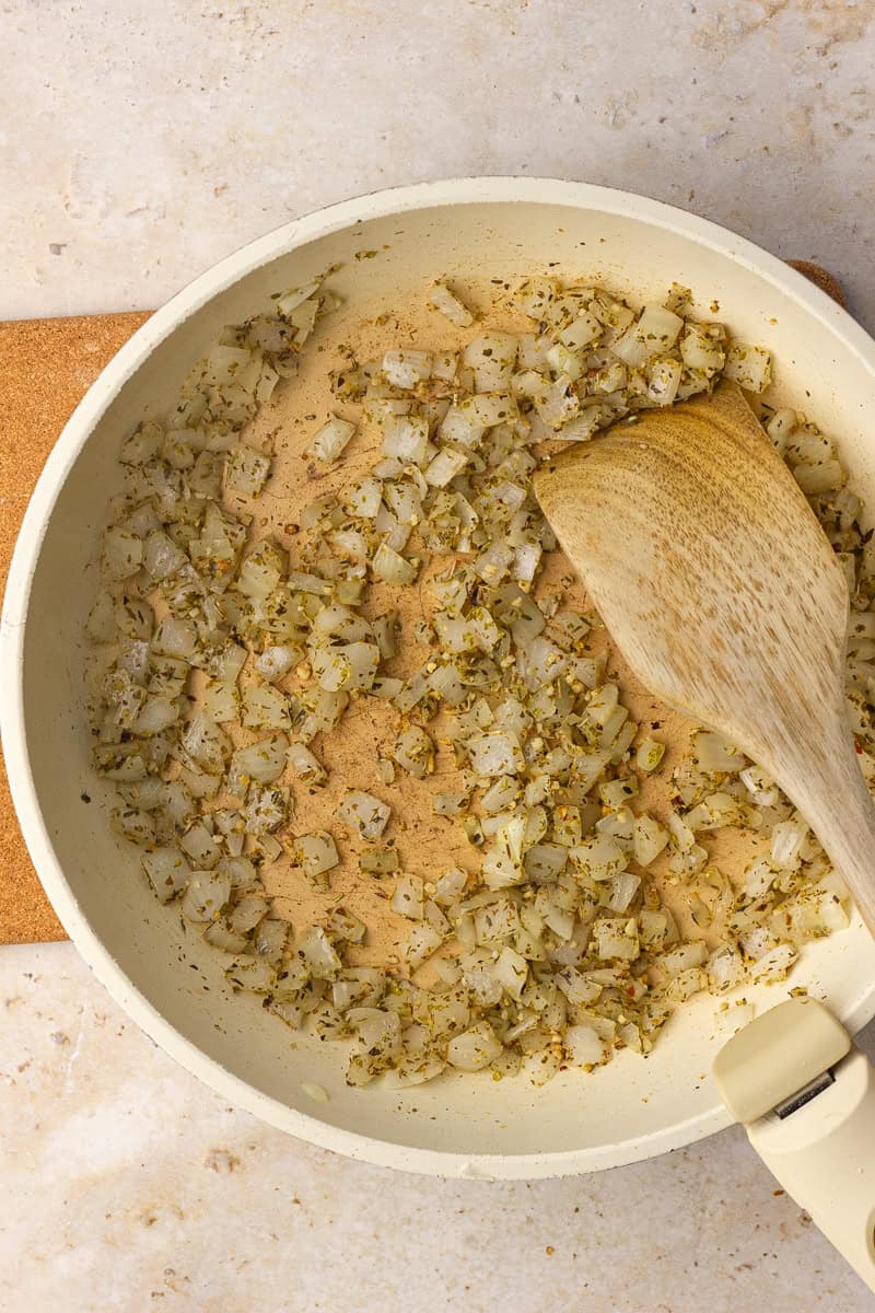Diced onions sautéing with Italian seasoning in a light skillet with a wooden spatula.