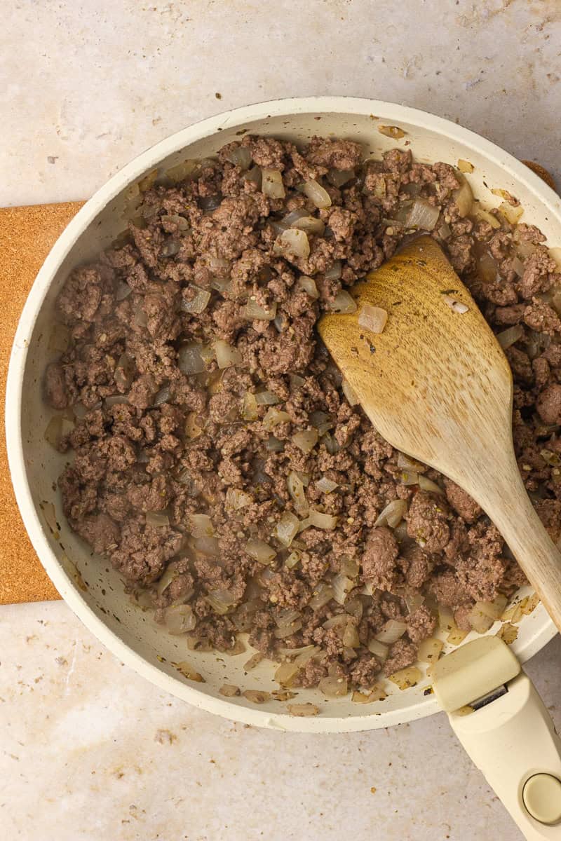 Ground beef browns with onions and herbs in a skillet, stirred with a wooden spatula.