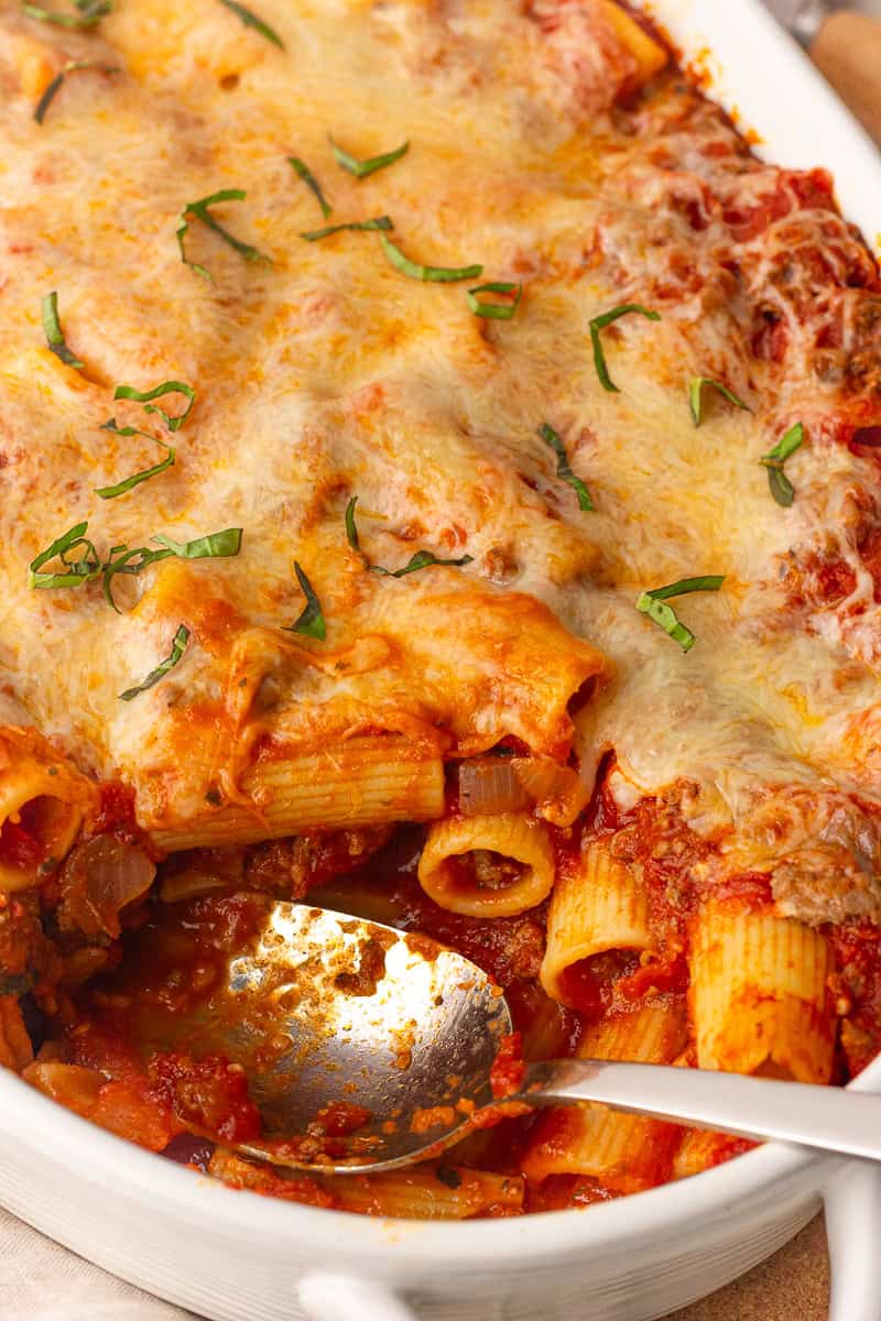 A spoon scoops into the cheesy baked rigatoni, revealing saucy noodles underneath.