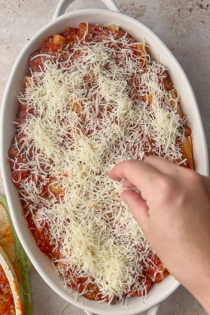 Hand sprinkles shredded mozzarella over sauced rigatoni al forno before baking.