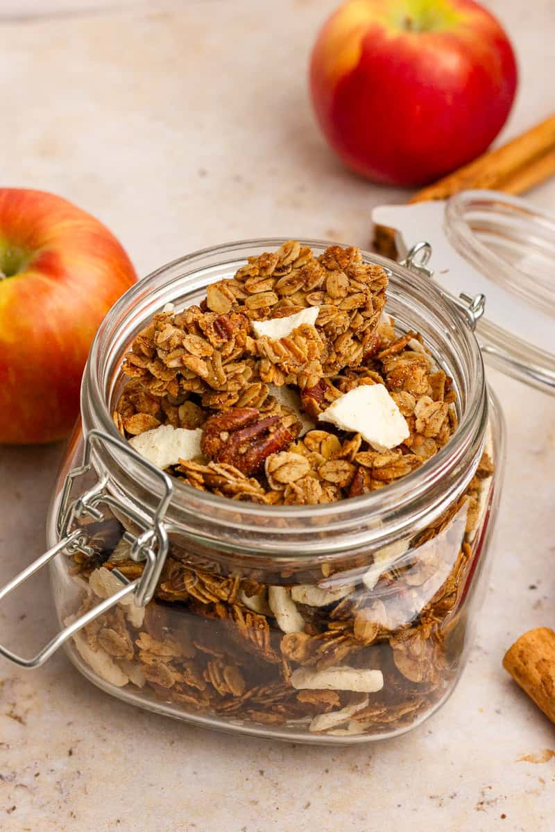 A glass jar filled with golden apple crumble granola, surrounded by fresh apples and cinnamon sticks.