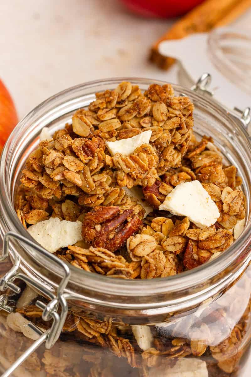 Close-up of crunchy apple crumble granola with oats, pecans, and apple pieces in a clear jar.