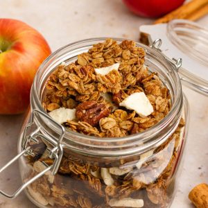 Jar of homemade apple crumble granola with visible pecans and apple pieces on a neutral background.