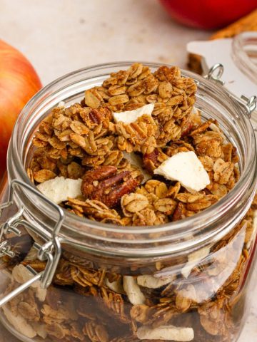 Jar of homemade apple crumble granola with visible pecans and apple pieces on a neutral background.
