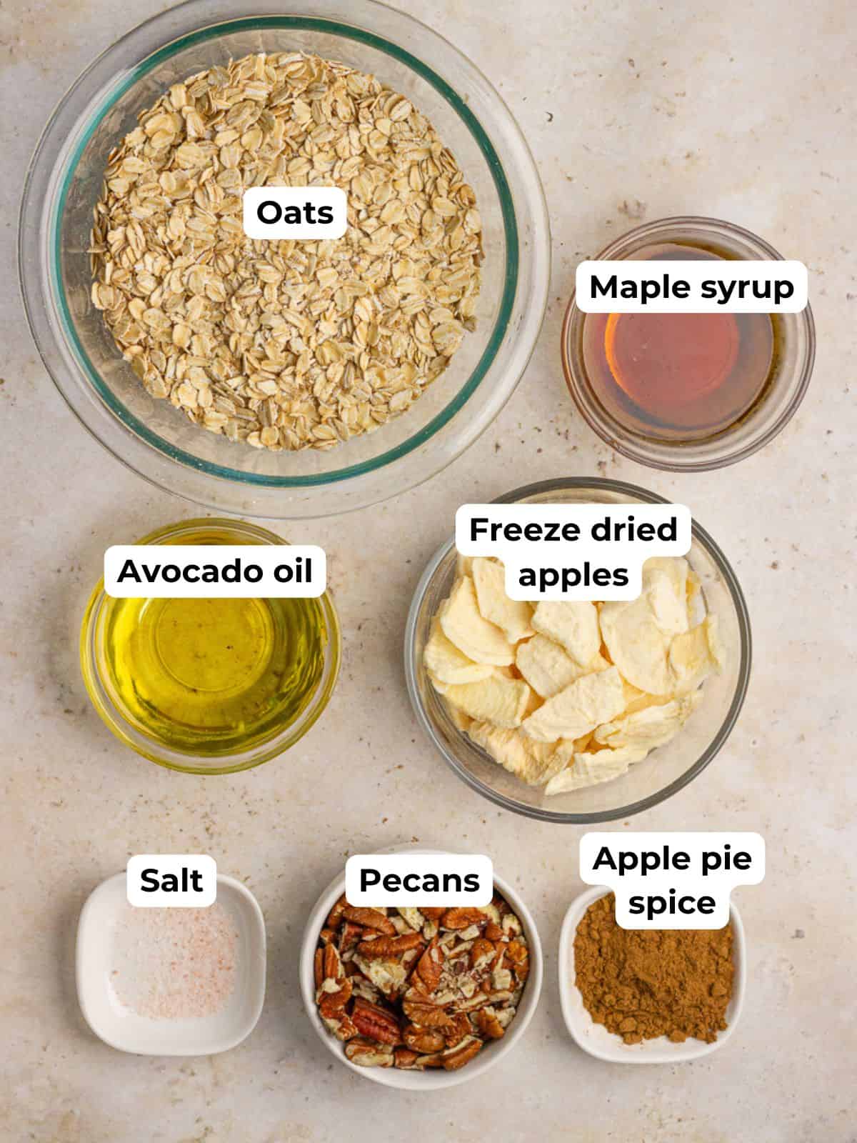 Overhead layout of apple crumble granola ingredients labeled in small bowls, including oats, pecans, and apple pie spice.