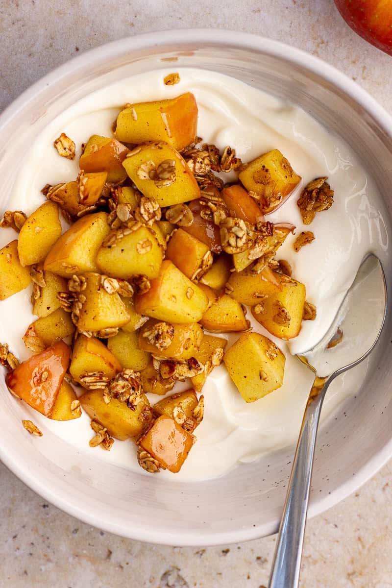 Bowl of apple pie yogurt topped with warm cinnamon apples and granola, ready to eat with a spoon.