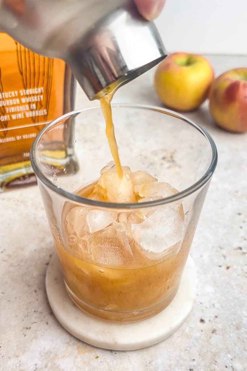 A cocktail shaker is pouring the apple whiskey mixture over ice into a glass.