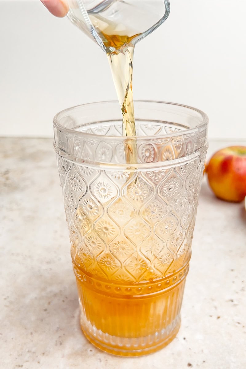 Whiskey is being poured from a small glass into a larger mixing glass with apple cider and apple butter.