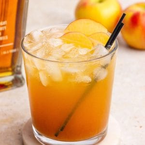 A finished apple whiskey cocktail with apple slice garnish sits on a marble coaster with apples and whiskey nearby.