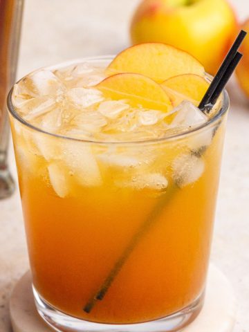A finished apple whiskey cocktail with apple slice garnish sits on a marble coaster with apples and whiskey nearby.