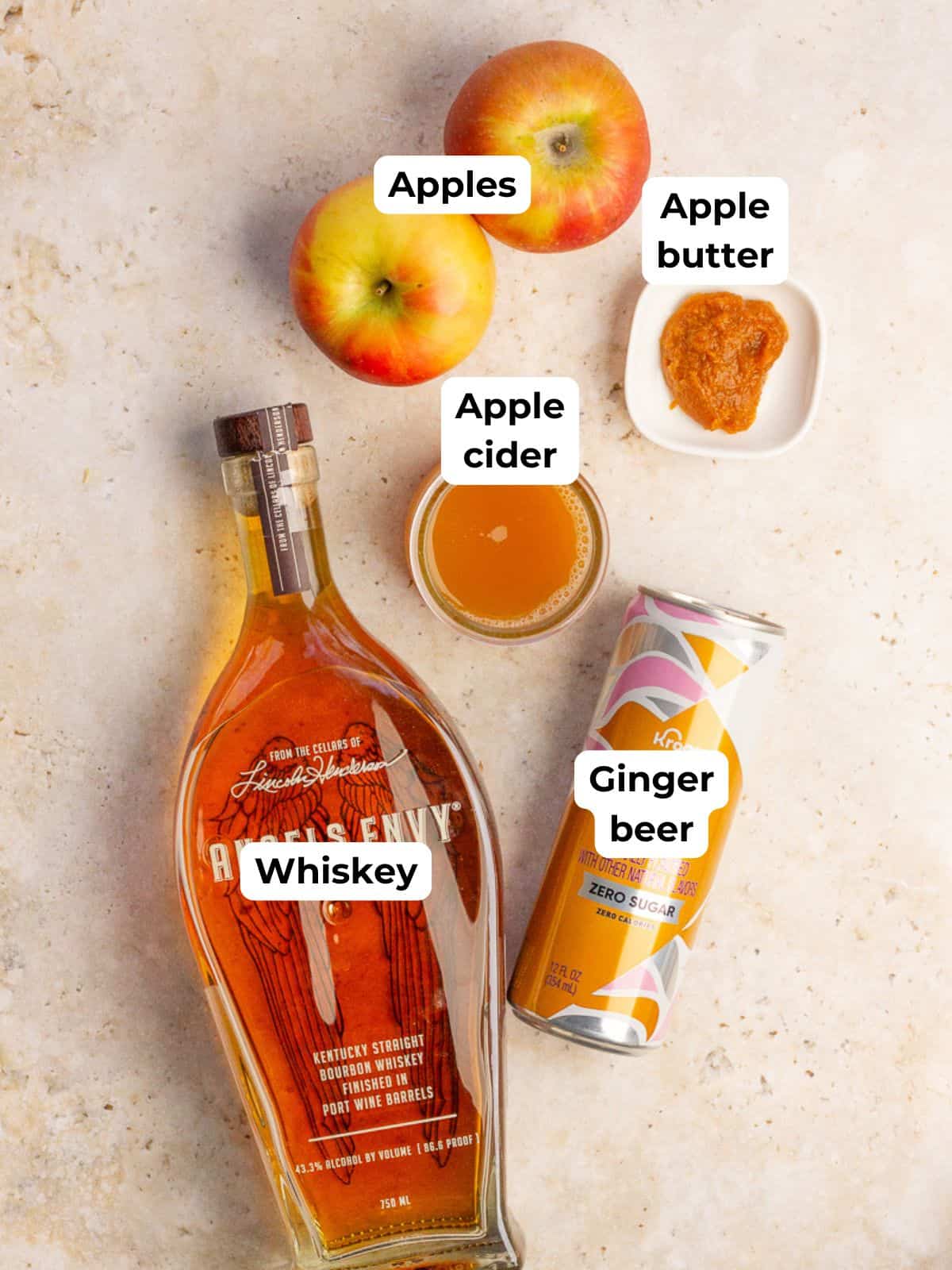 Ingredients for the apple whiskey cocktail, including whiskey, apples, apple cider, apple butter, and ginger beer, are displayed on a countertop.