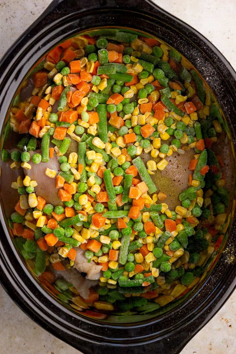 Frozen mixed vegetables are spread over chicken in the slow cooker.