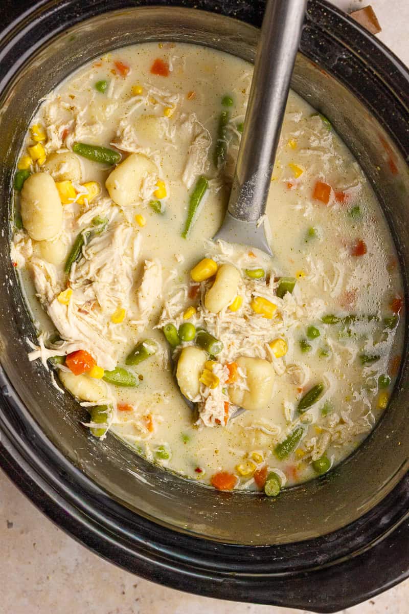 The slow cooker is filled with creamy chicken pot pie gnocchi soup and a ladle ready to serve.
