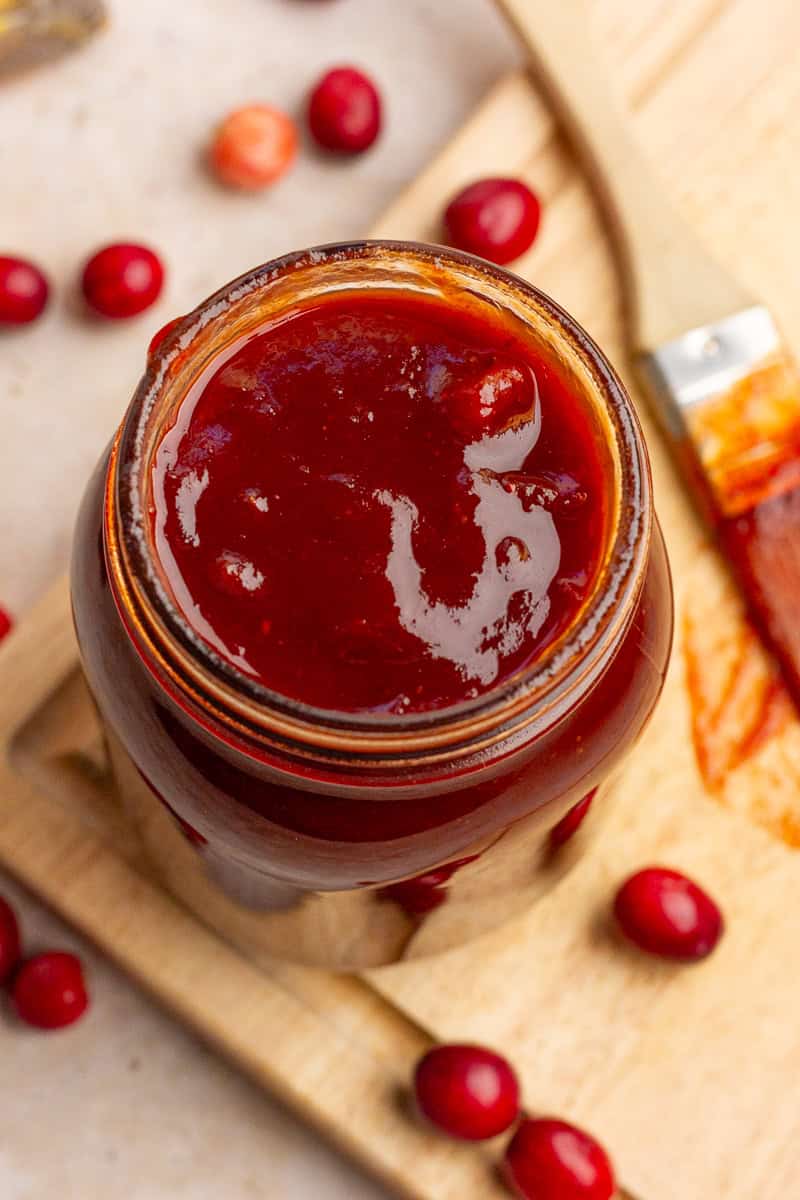 A jar of glossy cranberry BBQ sauce sits on a wooden board with fresh cranberries scattered around it.