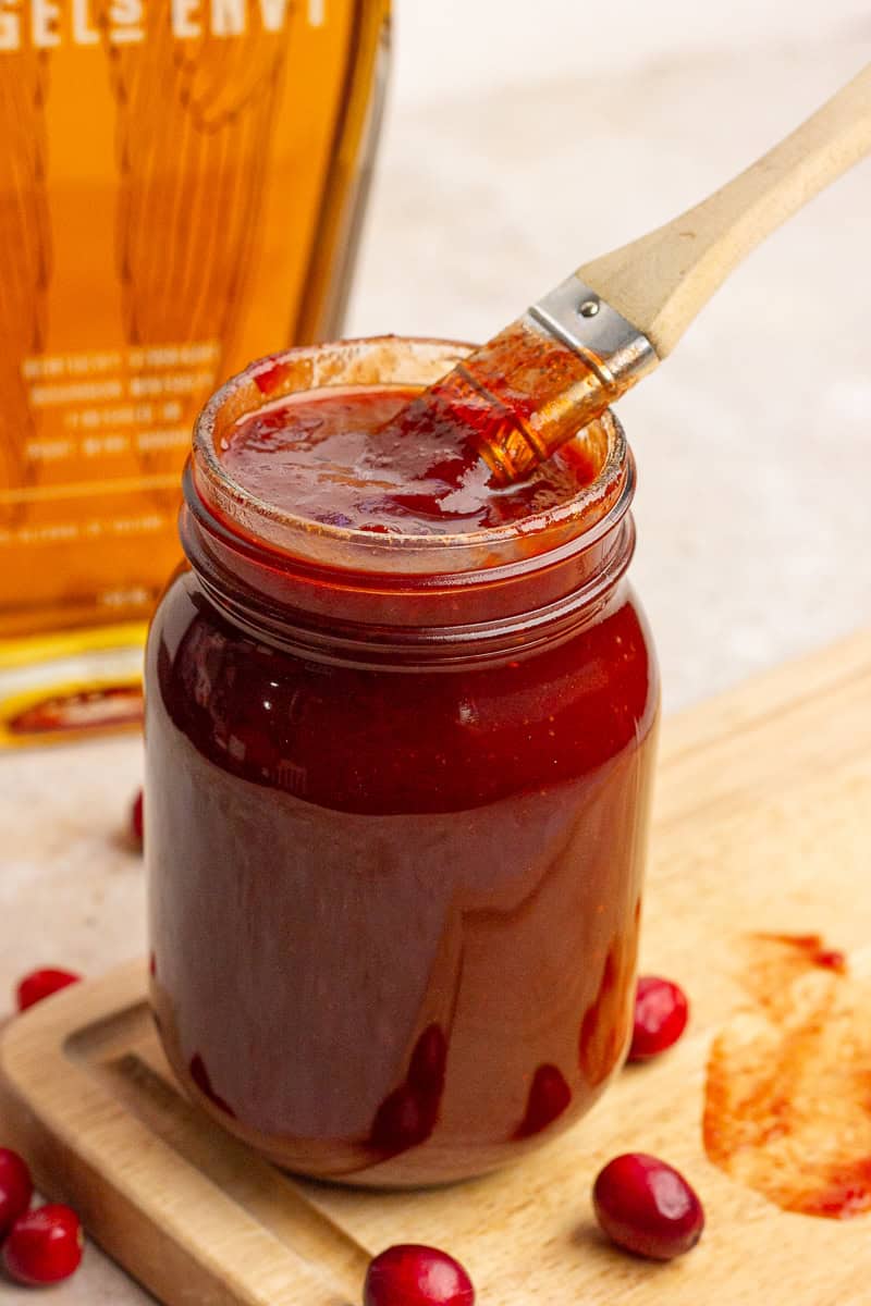 A jar of cranberry BBQ sauce is shown with a basting brush resting on top of it.