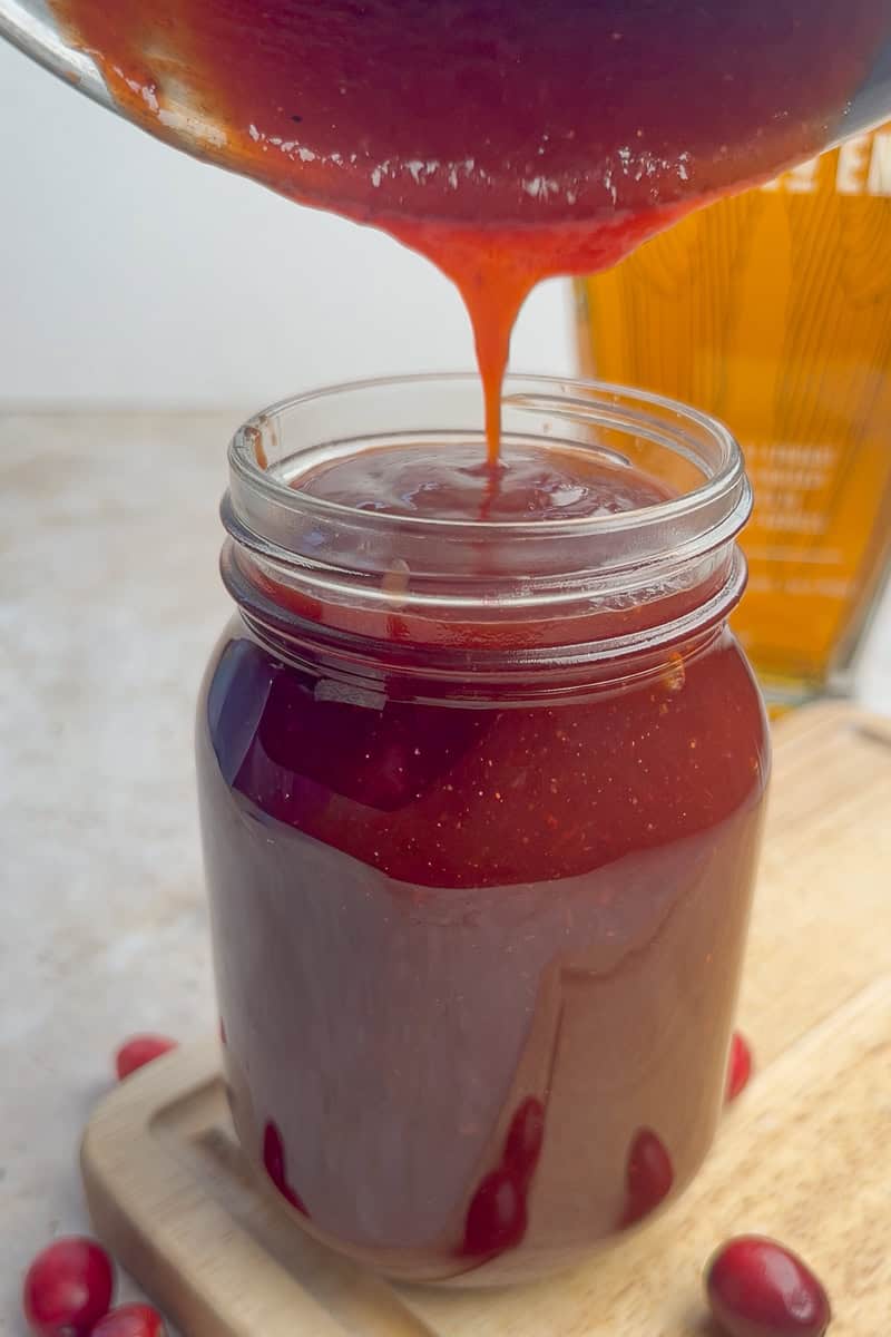 Warm cranberry BBQ sauce is being poured into a glass jar from a saucepan.