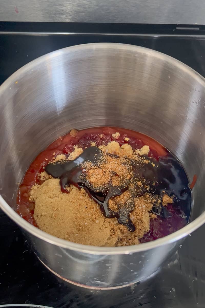 A saucepan filled with ketchup, cranberry sauce, brown sugar, molasses, and seasoning sits on a stovetop before stirring.