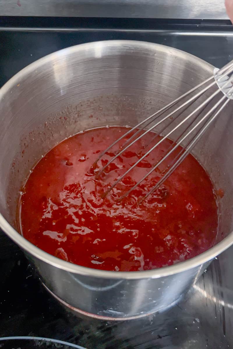 A whisk stirs a pot of simmering cranberry BBQ sauce on the stove.