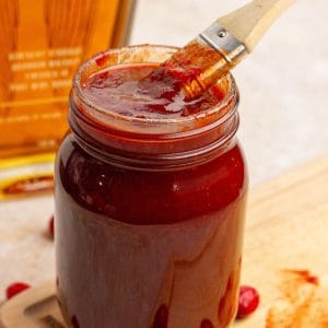 A jar of cranberry BBQ sauce is shown with a brush dipped inside and fresh cranberries around it.