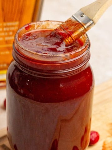 A jar of cranberry BBQ sauce is shown with a brush dipped inside and fresh cranberries around it.