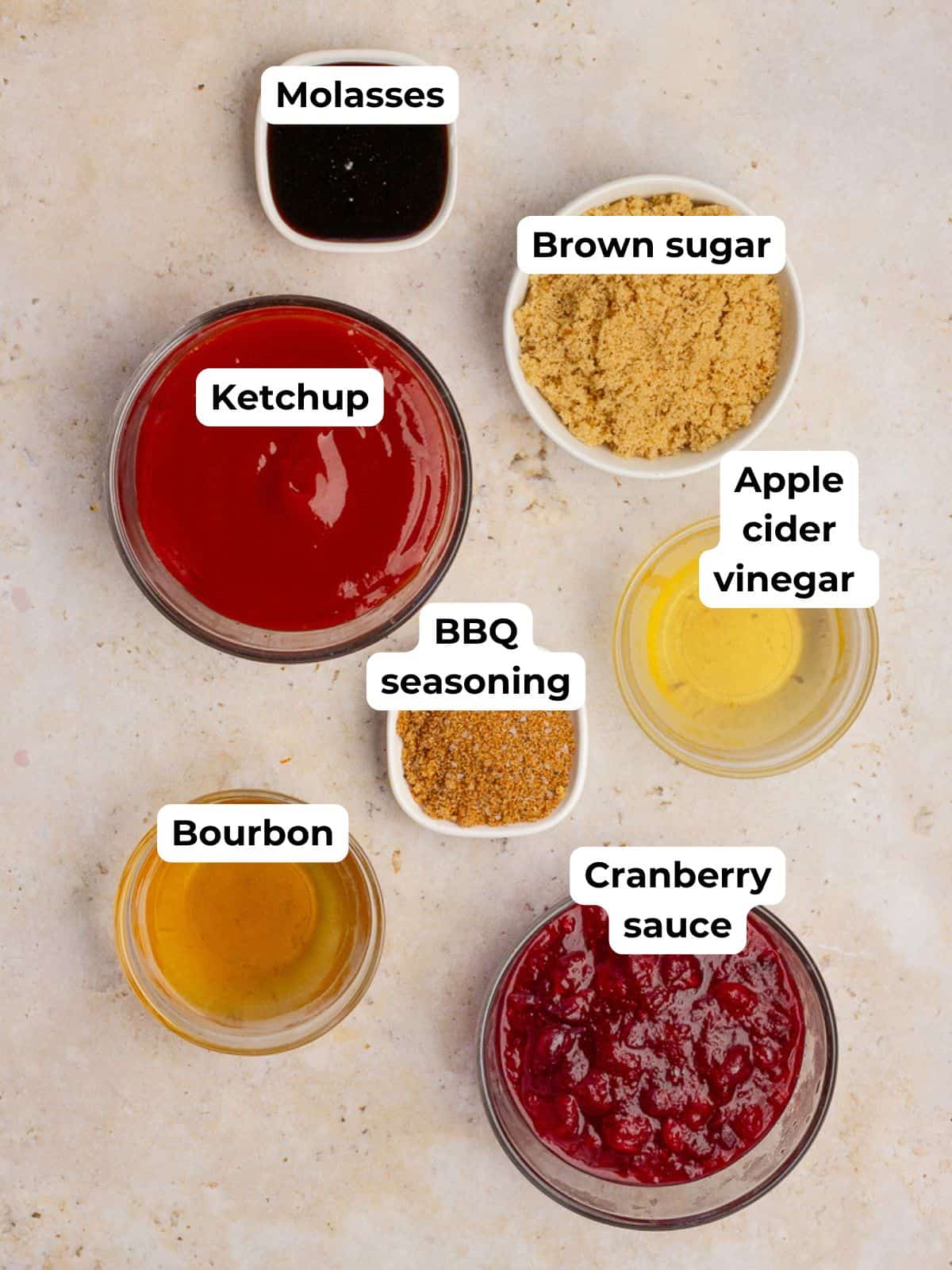 Individual bowls of ketchup, cranberry sauce, brown sugar, molasses, vinegar, bourbon, and BBQ seasoning are arranged on a countertop.