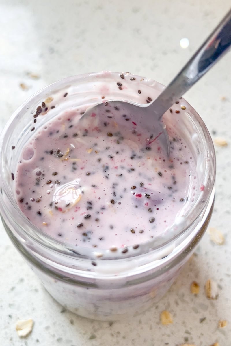 A spoon rests in an open jar of cranberry overnight oats, showing the creamy mixture with chia seeds.