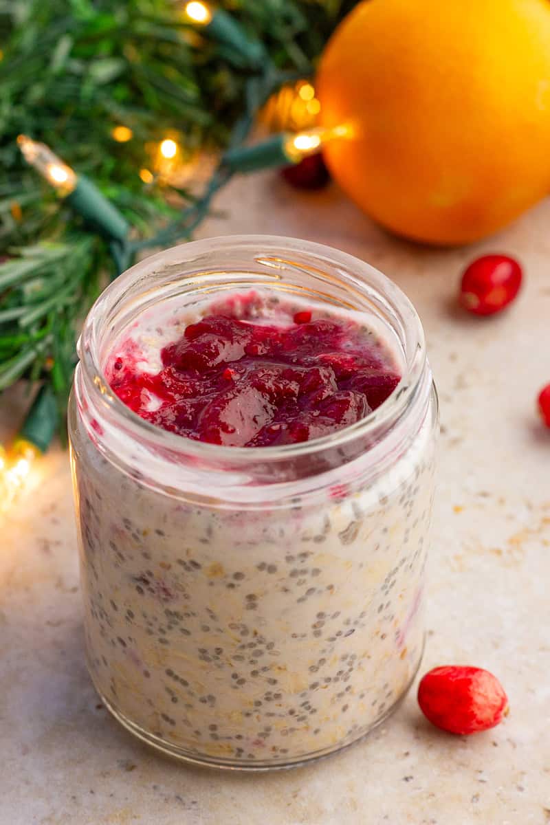 A jar of cranberry overnight oats sits on a counter with holiday lights and an orange in the background.