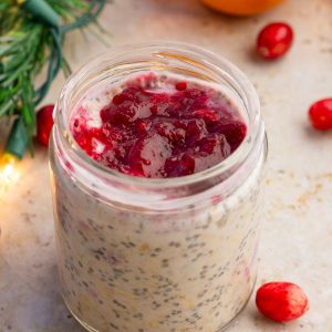 A jar of cranberry overnight oats is topped with cranberry sauce and surrounded by fresh cranberries and holiday greenery.