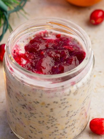 A jar of cranberry overnight oats is topped with cranberry sauce and surrounded by fresh cranberries and holiday greenery.