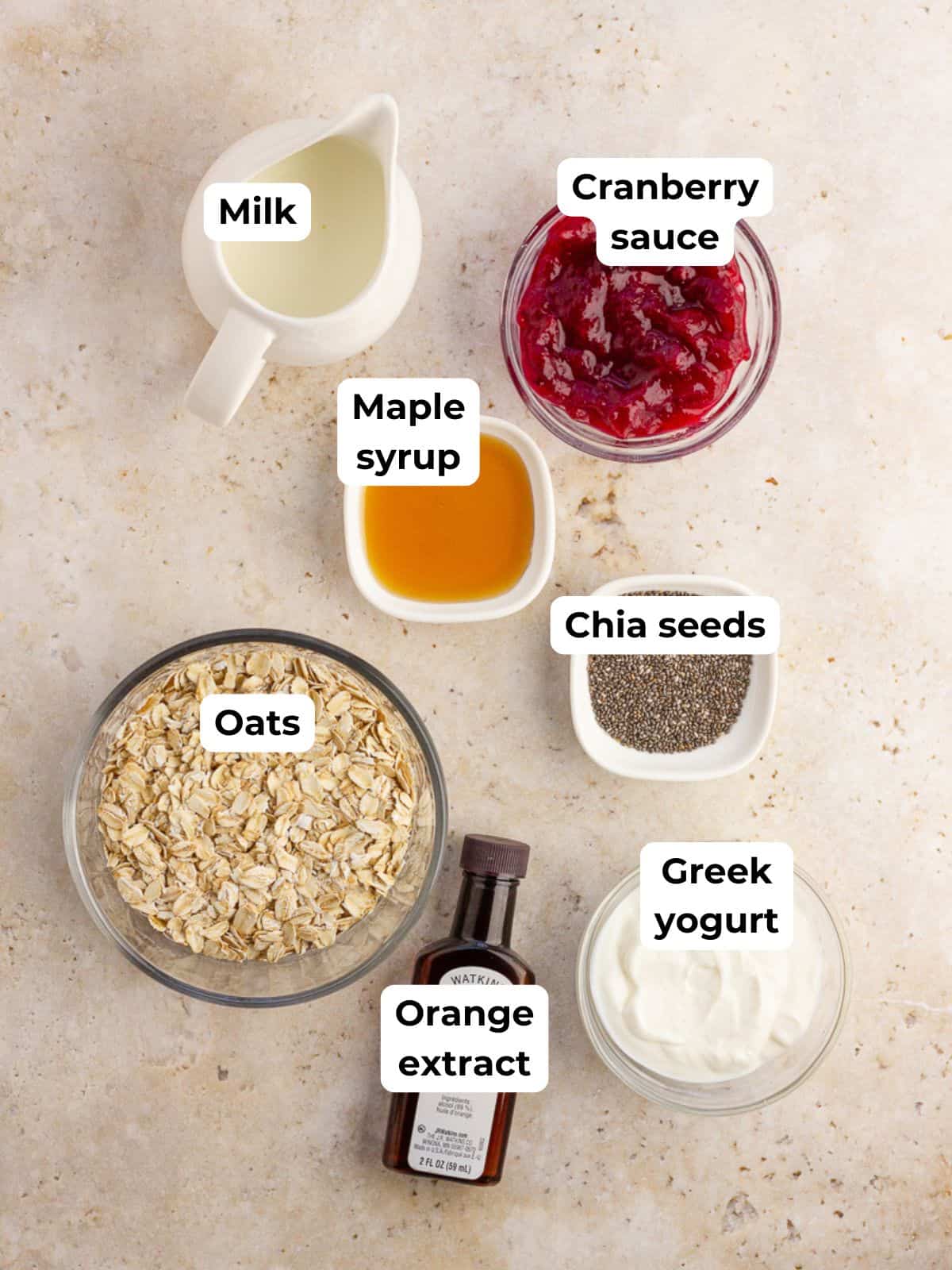 A flat lay displays ingredients for cranberry overnight oats, including oats, milk, yogurt, chia seeds, maple syrup, orange extract, and cranberry sauce.