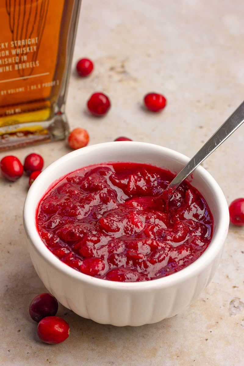 A white bowl of chunky cranberry sauce sits beside scattered fresh cranberries and a bottle of bourbon.