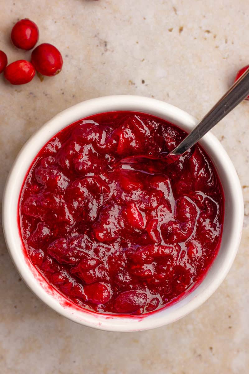 A top-down view shows a white bowl filled with thick, glossy cranberry sauce with visible whole berries.