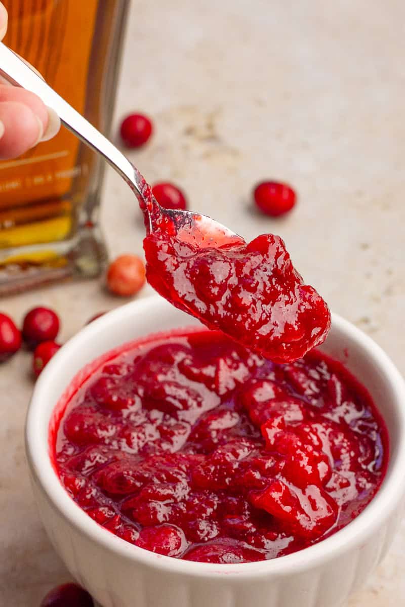 A spoon lifts a scoop of chunky cranberry sauce from the bowl with fresh cranberries and bourbon in the background.