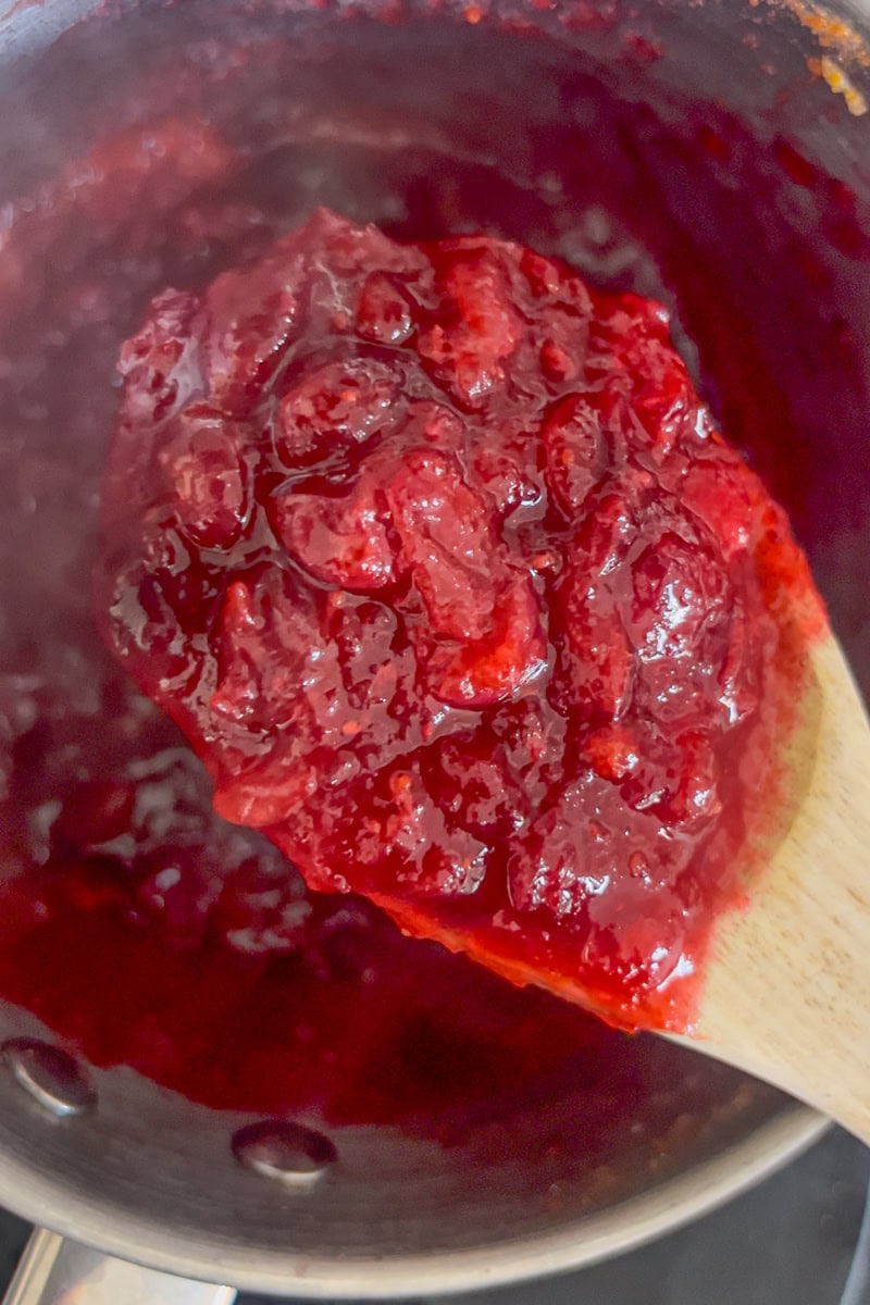 A wooden spoon holds thick, simmering cranberry sauce over a pot on the stovetop.