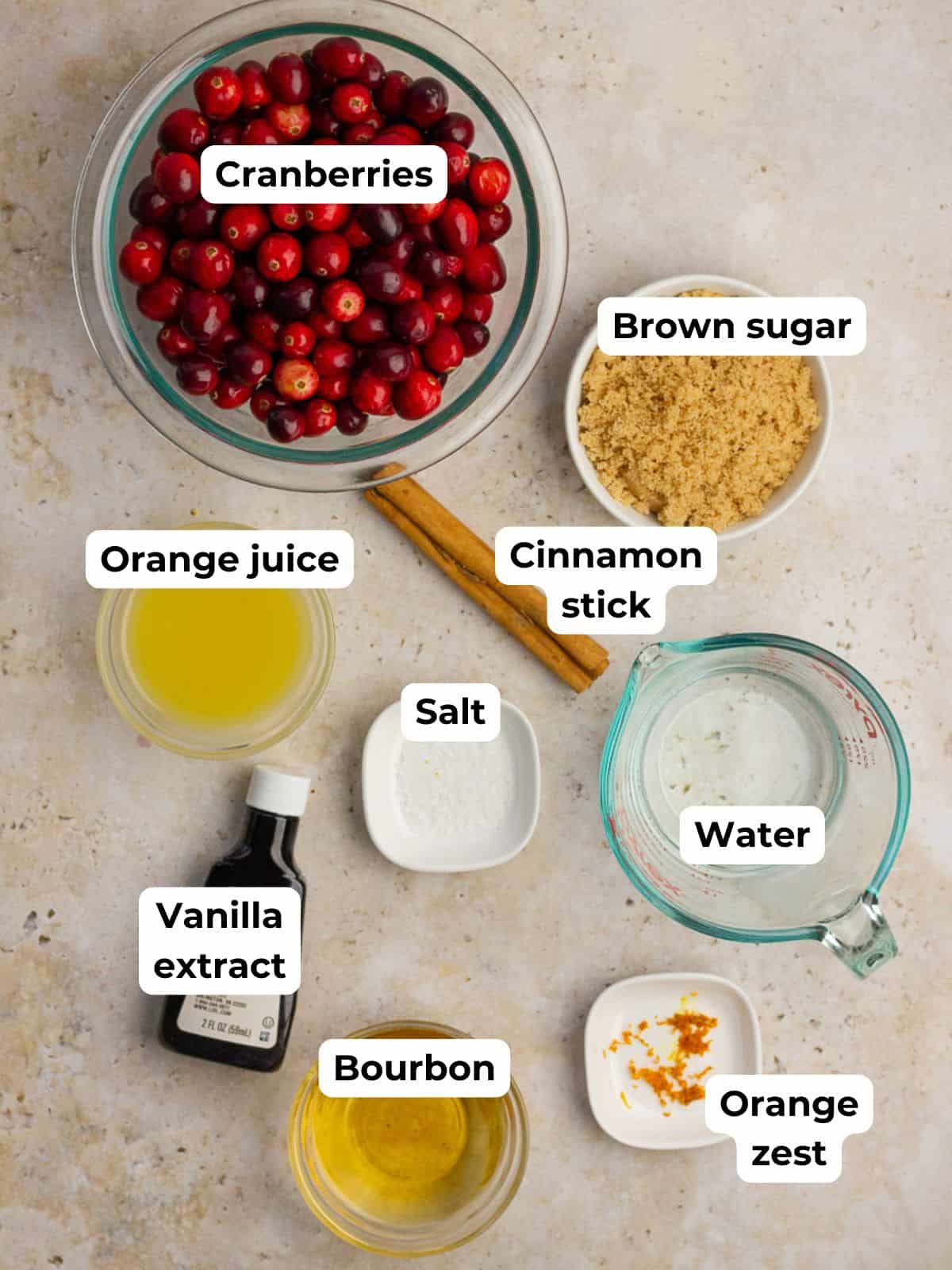 Labeled ingredients including cranberries, brown sugar, orange juice, cinnamon, vanilla, water, salt, bourbon, and orange zest are arranged on a countertop.