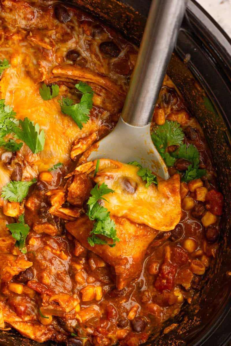 Serving spoon lifting a cheesy scoop of crockpot chicken enchilada casserole.