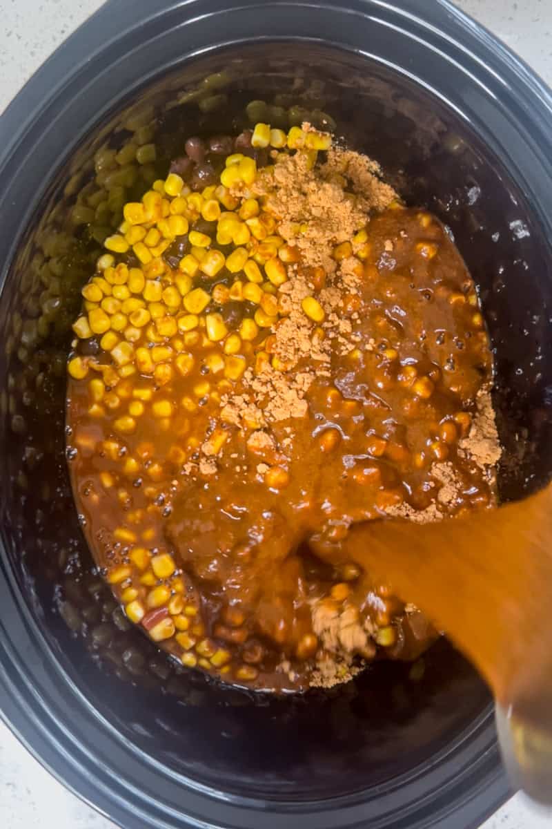 Enchilada sauce being poured over corn, beans, and taco seasoning in a crockpot.