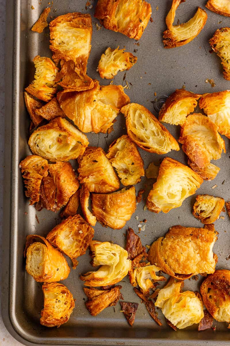 Golden baked croissant croutons are scattered across a sheet pan showing their crispy, flaky layers.