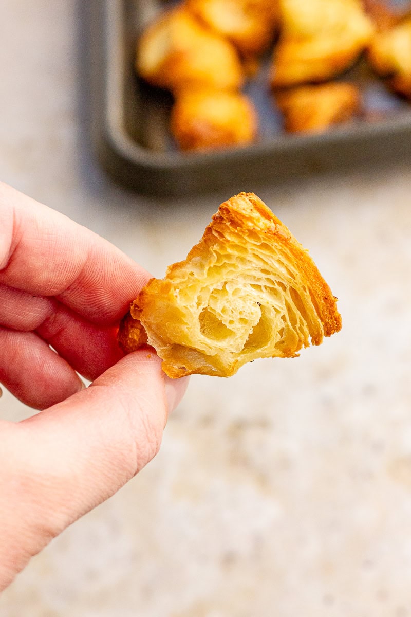 A hand holds a single croissant crouton to show its flaky, layered texture up close.