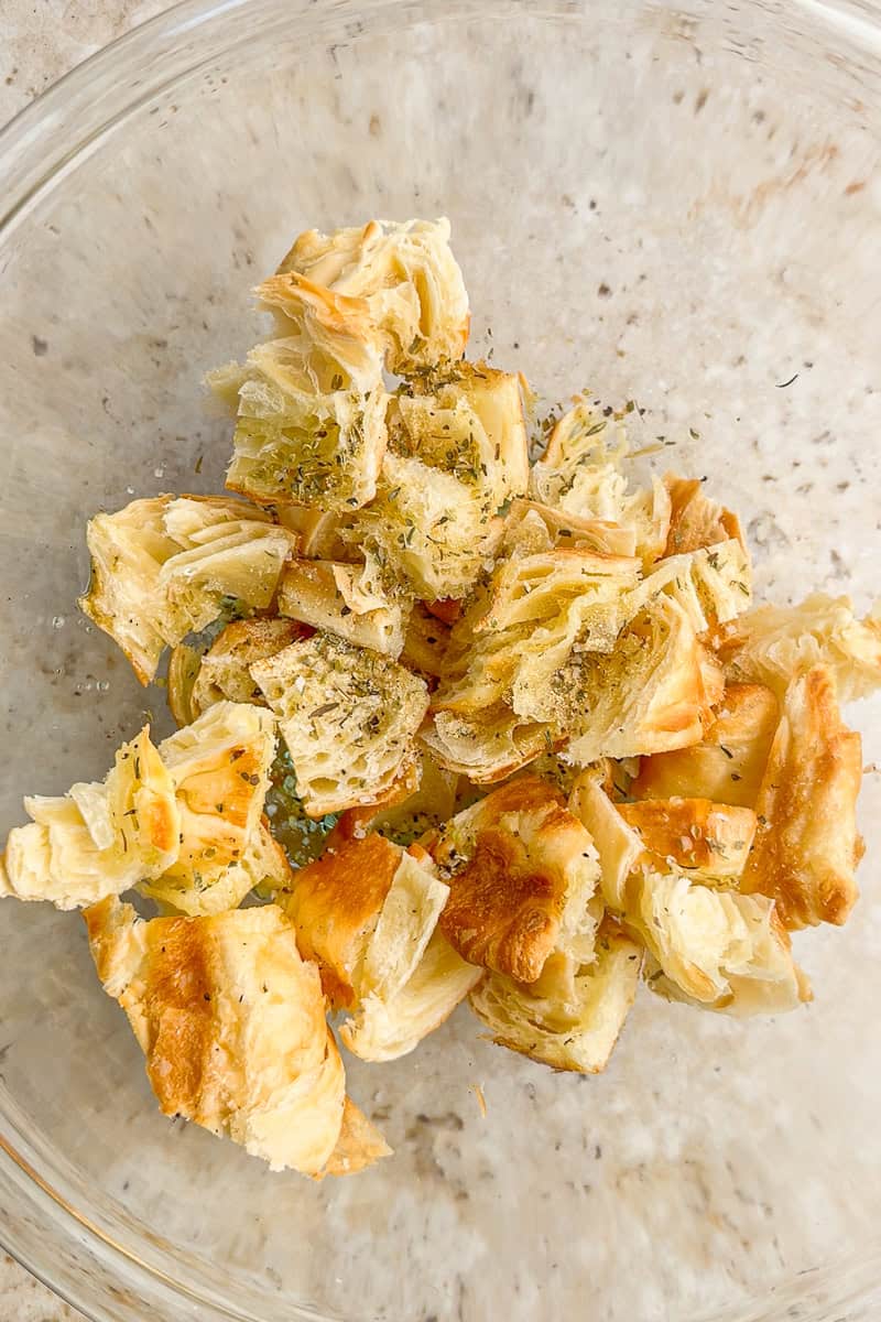 Croissant pieces tossed with olive oil and seasonings sit in a glass bowl ready for baking.