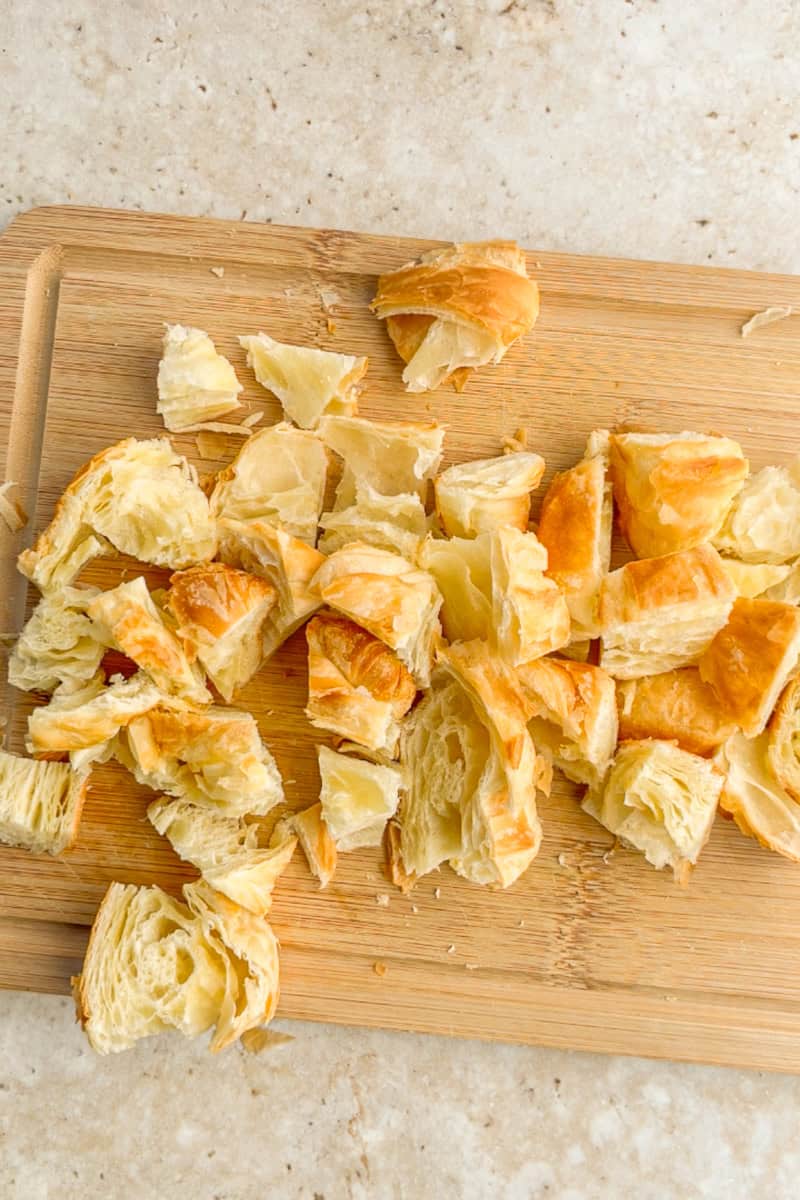 Chopped croissant pieces rest on a wooden cutting board before being seasoned and baked.