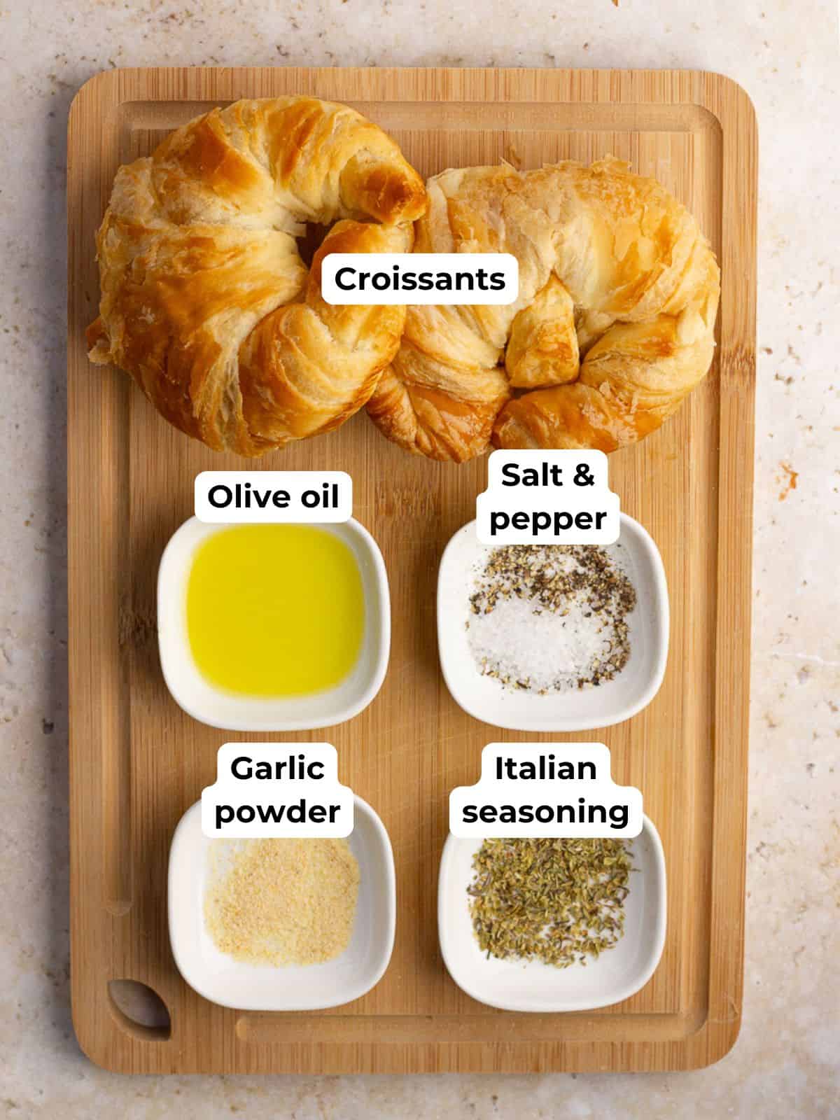 Croissants, olive oil, garlic powder, Italian seasoning, and salt and pepper are arranged on a small wooden board.