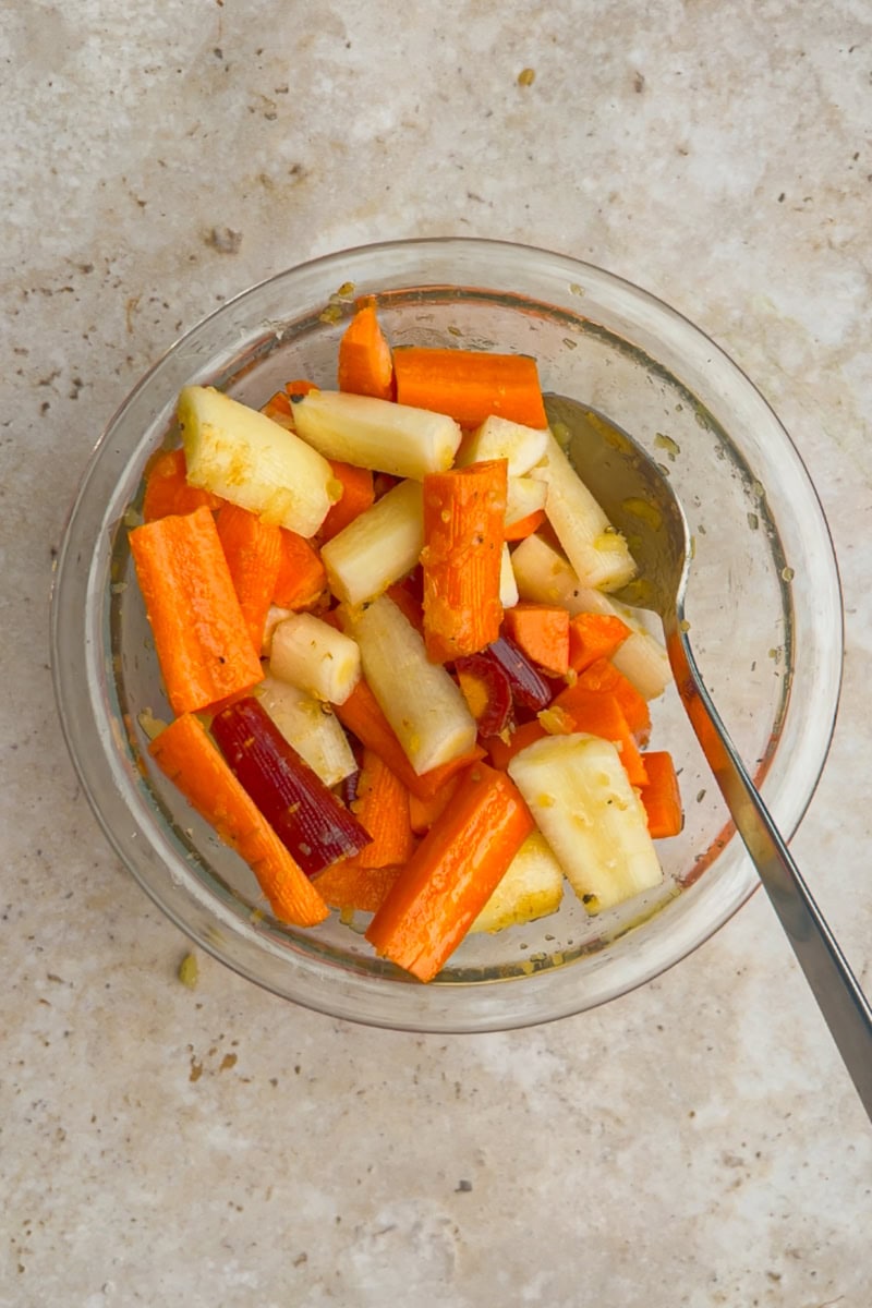 A glass bowl filled with chopped rainbow carrots tossed in a maple bourbon glaze sits on a light countertop.